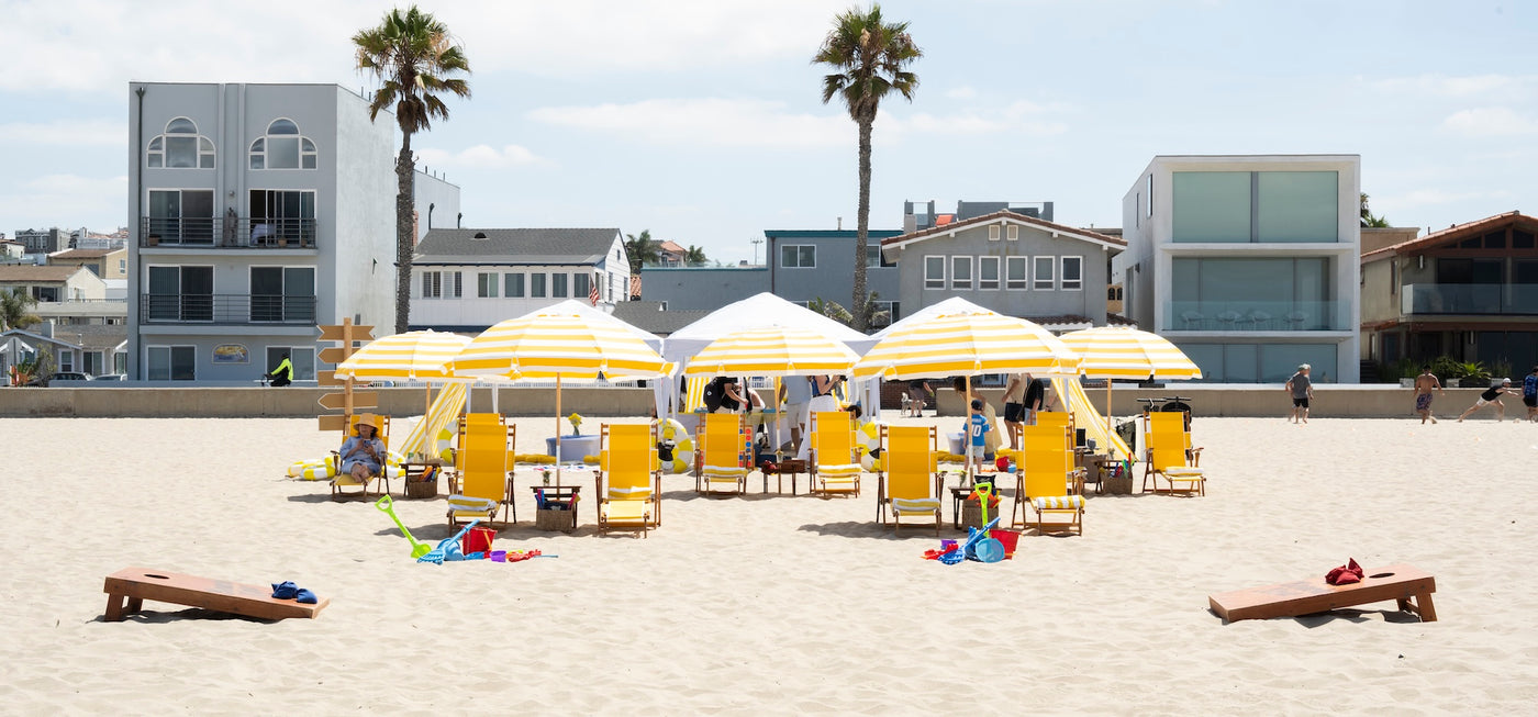 Vibrant Beach Now custom party setup on a white-sand beach with yellow striped umbrellas, comfy lounge chairs, sand toys, and outdoor games.